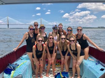Group of women in matching black tank tops smiling on a leisure boat with a cable-stayed bridge, blue sky and coastal waters on a sunny summer day.