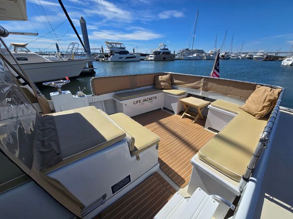 Sunlit yacht cockpit with tan cushioned seating, teak-style deck and small table, American flag flying as yachts and sailboats line a busy marina under a blue sky