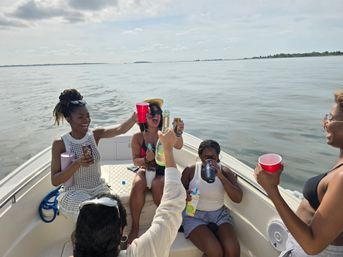 Friends laughing and toasting with cups and bottles on the bow of a small boat during a sunny coastal boat outing on calm waters.