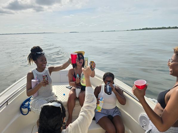 Friends laughing and toasting with cups and bottles on the bow of a small boat during a sunny coastal boat outing on calm waters.