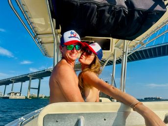 Smiling shirtless man and woman in sunglasses wearing matching red, white and blue trucker hats embrace and kiss while sitting on a boat under a large bridge over bright blue water on a sunny day.