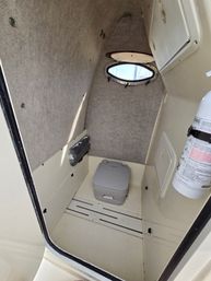 Compact boat head cabin with a gray portable marine toilet on a molded deck, oval porthole letting in daylight, carpeted walls, small storage hatches and a mounted fire extinguisher.