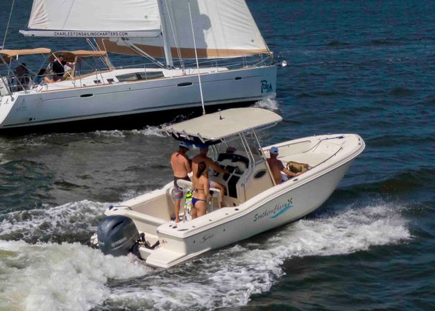 Group on a center-console motorboat in swimwear cruising past a white sailboat on open blue water, creating a foamy wake on a sunny day.