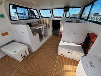 Bright motorboat pilothouse interior with striped bench seating and folding table, white coolers, life ring and fire extinguisher on faux‑teak deck, large windows revealing marina dock slips