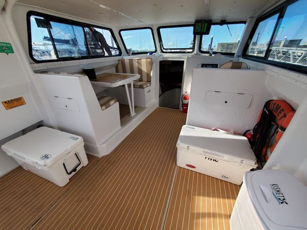 Bright motorboat pilothouse interior with striped bench seating and folding table, white coolers, life ring and fire extinguisher on faux‑teak deck, large windows revealing marina dock slips