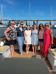 Six women smiling together barefoot on a boat deck at a sunny marina, with sailboats and a harbor bridge in the blue-sky background
