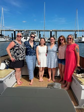 Six women smiling together barefoot on a boat deck at a sunny marina, with sailboats and a harbor bridge in the blue-sky background