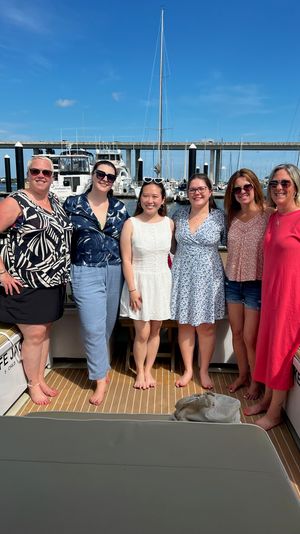 Six women smiling together barefoot on a boat deck at a sunny marina, with sailboats and a harbor bridge in the blue-sky background