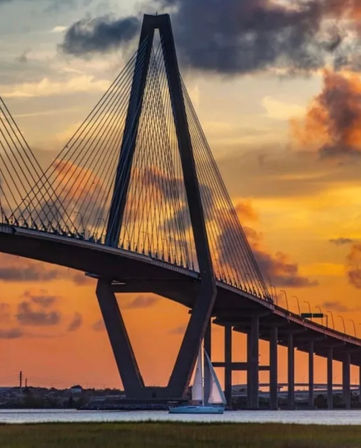 Cable-stayed bridge silhouetted against a vibrant orange sunset over coastal waters, a sailboat gliding beneath the span.
