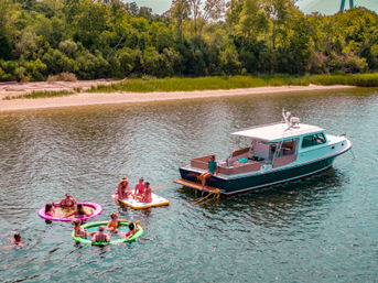 Group of people on colorful inflatable floats and a paddleboard tethered to a moored motorboat near a sandy, tree-lined riverbank on a sunny summer day.