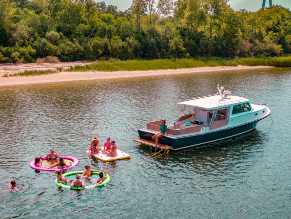 Group of people on colorful inflatable floats and a paddleboard tethered to a moored motorboat near a sandy, tree-lined riverbank on a sunny summer day.