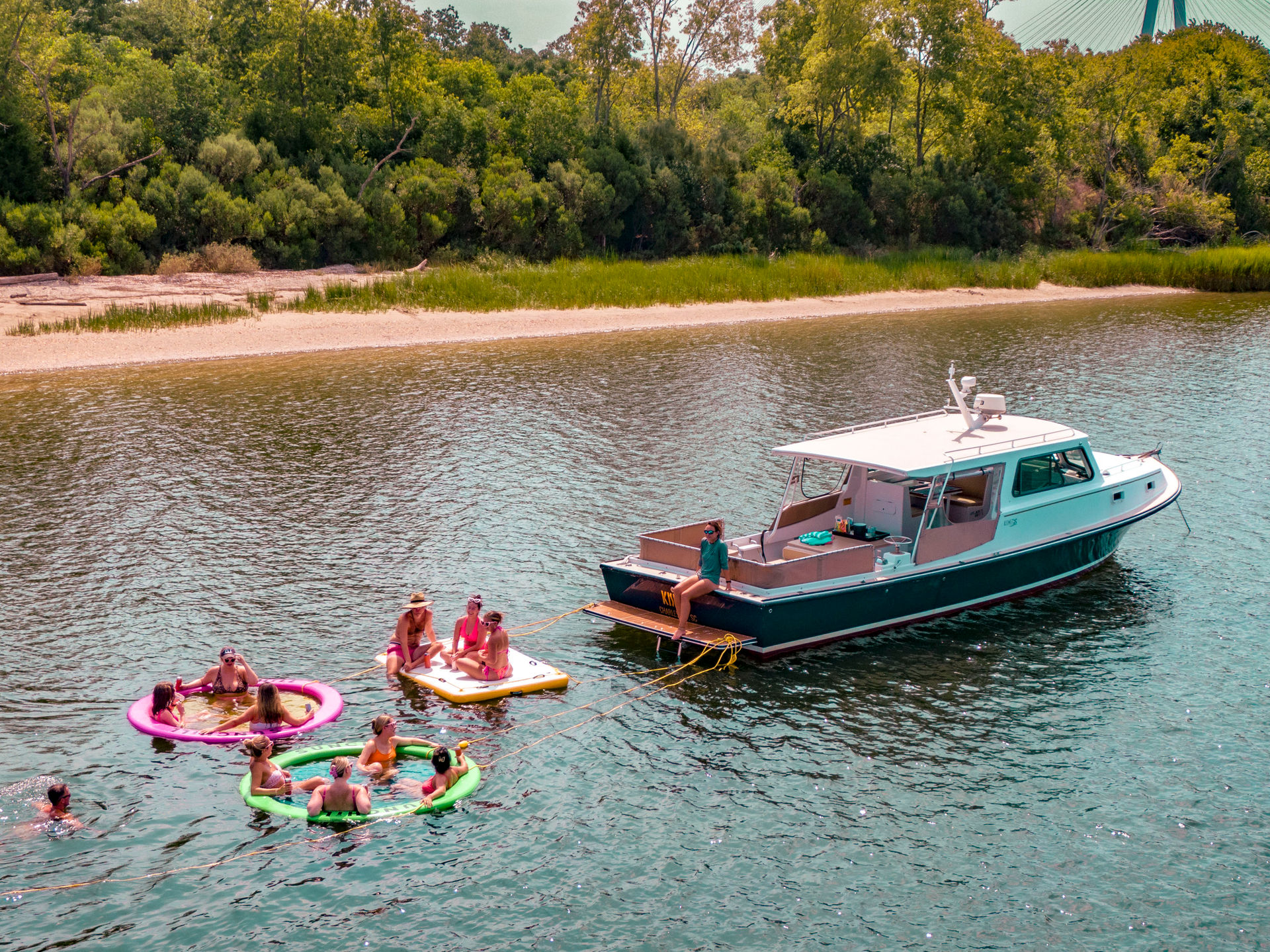 Group of people on colorful inflatable floats and a paddleboard tethered to a moored motorboat near a sandy, tree-lined riverbank on a sunny summer day.
