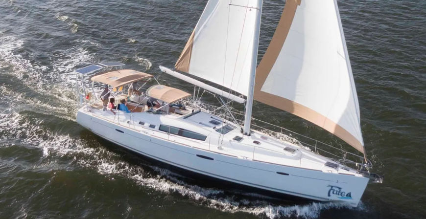 Aerial view of a white cruising sailboat with tan-accented sails and shaded cockpit, speeding through choppy coastal waters.