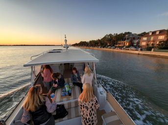 Group of friends on a small motorboat cruising a residential waterfront canal at sunset, passing seaside houses and calm water.