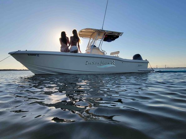 Two people sitting on the bow of a small center-console boat in calm coastal waters at sunset, with rippling water reflections and a clear sky.