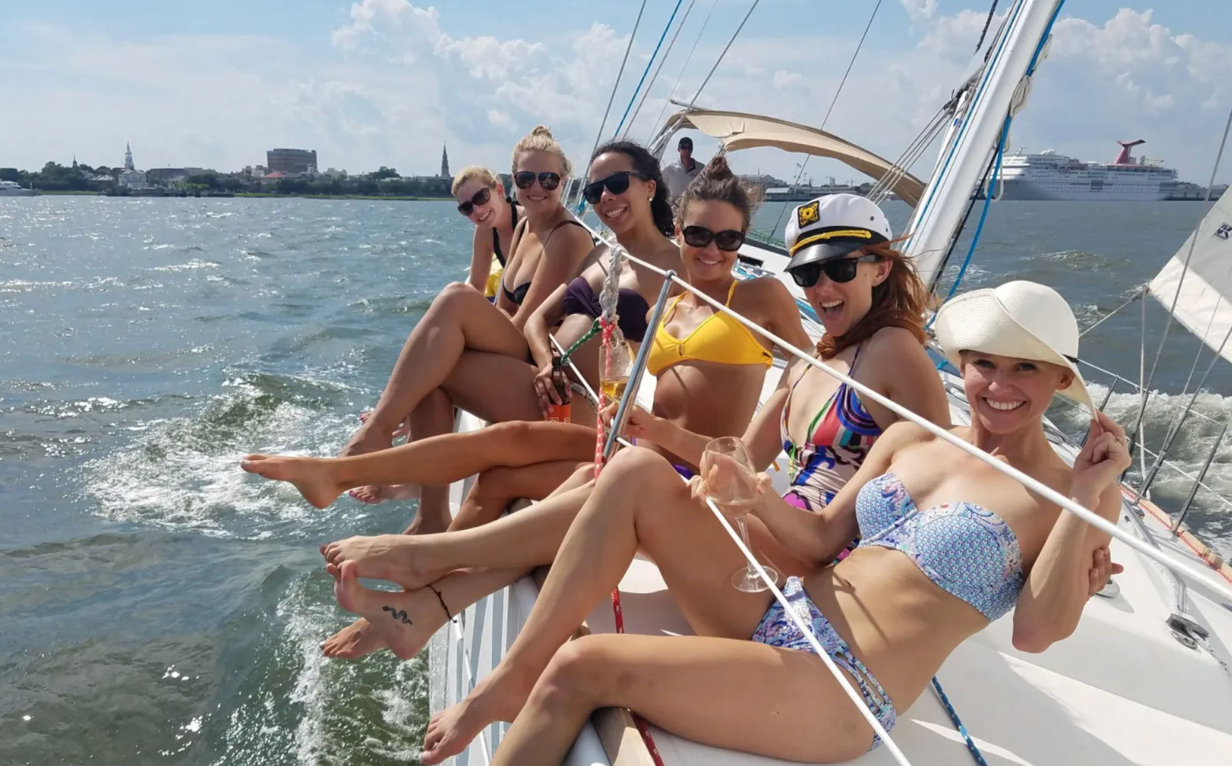Six women in colorful bikinis and sunglasses lounging on the rail of a sailing yacht, holding drinks and smiling on a sunny summer day with a coastal skyline and cruise ship in the harbor