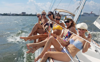Six women in colorful bikinis and sunglasses lounging on the rail of a sailing yacht, holding drinks and smiling on a sunny summer day with a coastal skyline and cruise ship in the harbor