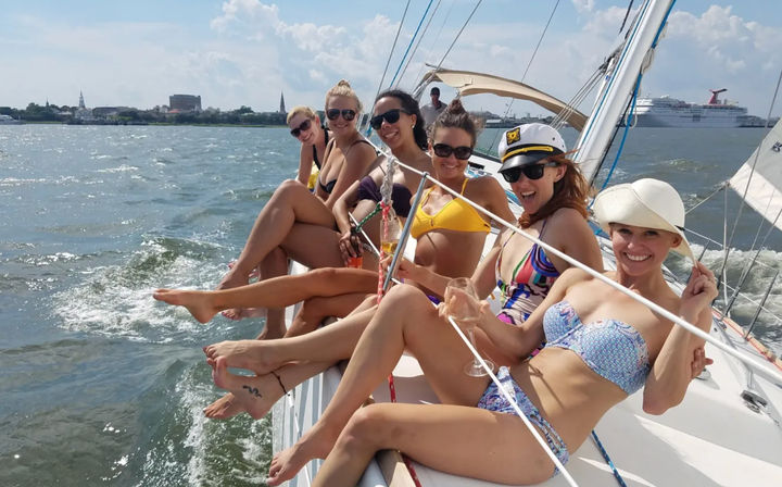 Six women in colorful bikinis and sunglasses lounging on the rail of a sailing yacht, holding drinks and smiling on a sunny summer day with a coastal skyline and cruise ship in the harbor