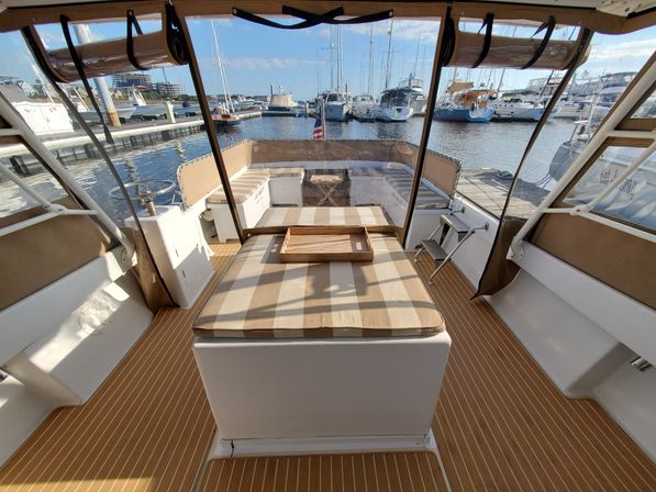 Sunlit yacht cockpit with striped cushioned sunpad and wooden tray on a teak-style deck, clear vinyl enclosure opening to a marina filled with moored sailboats and motorboats.