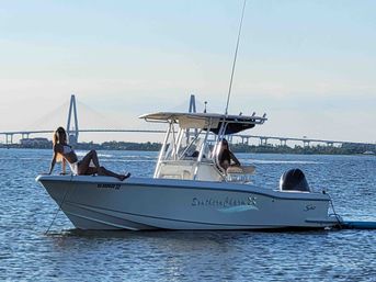 Two people lounging on a white center-console motorboat anchored in calm bay waters on a sunny afternoon, with a cable-stayed bridge and low shoreline in the background.