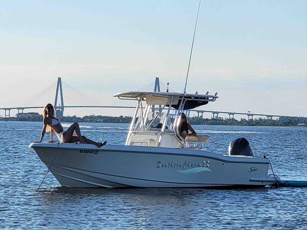 Two people lounging on a white center-console motorboat anchored in calm bay waters on a sunny afternoon, with a cable-stayed bridge and low shoreline in the background.