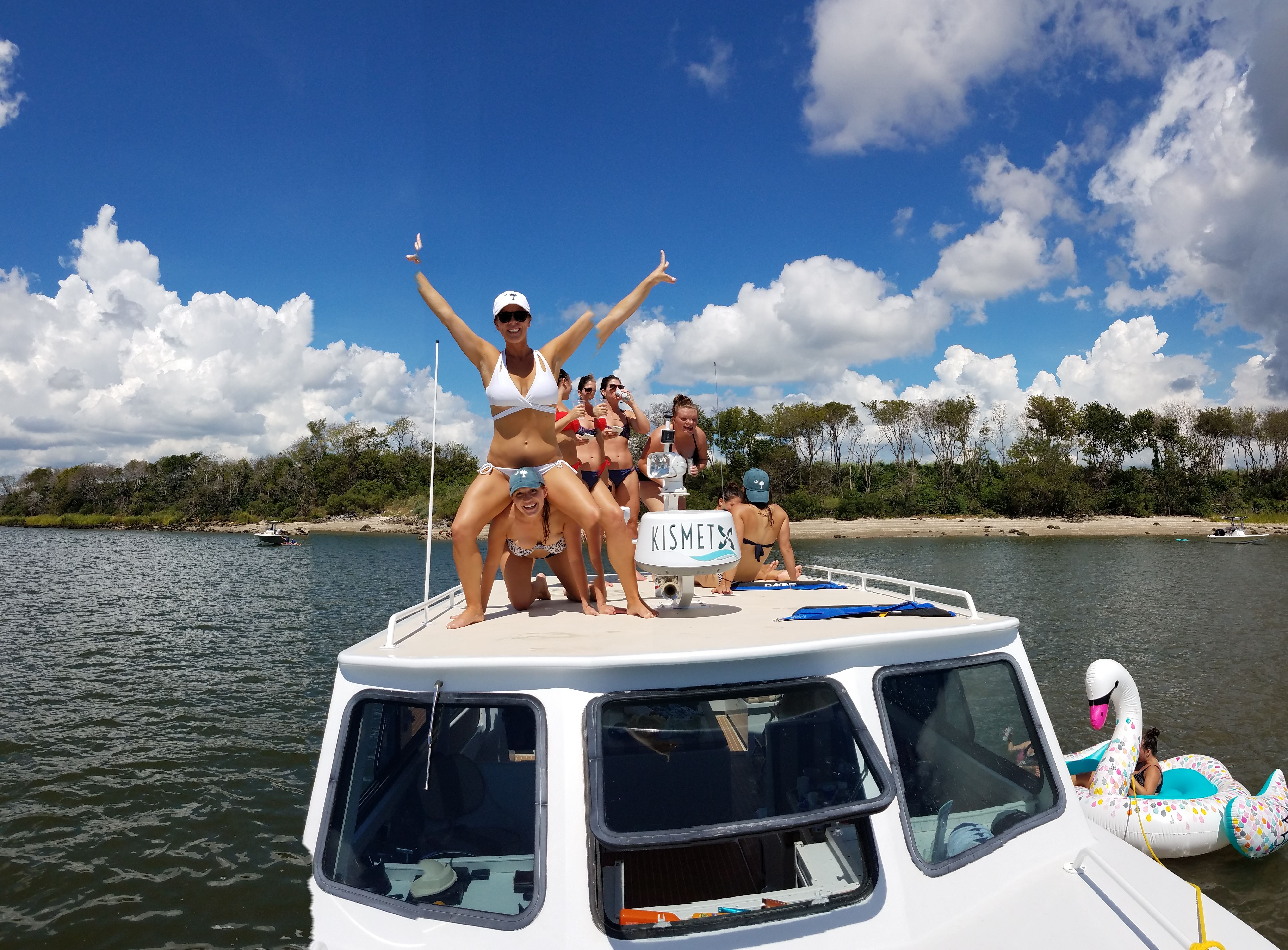 Group of friends in bikinis cheering on the bow of a small motorboat in a sunny coastal inlet, inflatable swan nearby, sandy shore and tree-lined shoreline under a bright blue sky with puffy clouds.