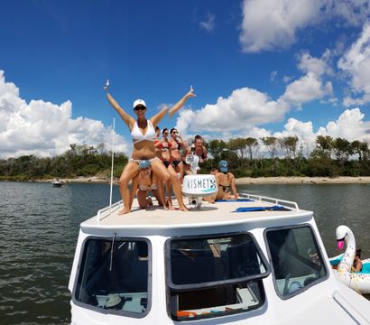 Group of friends in bikinis cheering on the bow of a small motorboat in a sunny coastal inlet, inflatable swan nearby, sandy shore and tree-lined shoreline under a bright blue sky with puffy clouds.