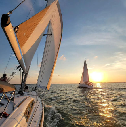 Two sailboats sailing on open water at sunset — close-up of white sails and deck with golden sunlight reflecting on rippling waves.