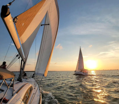 Two sailboats sailing on open water at sunset — close-up of white sails and deck with golden sunlight reflecting on rippling waves.