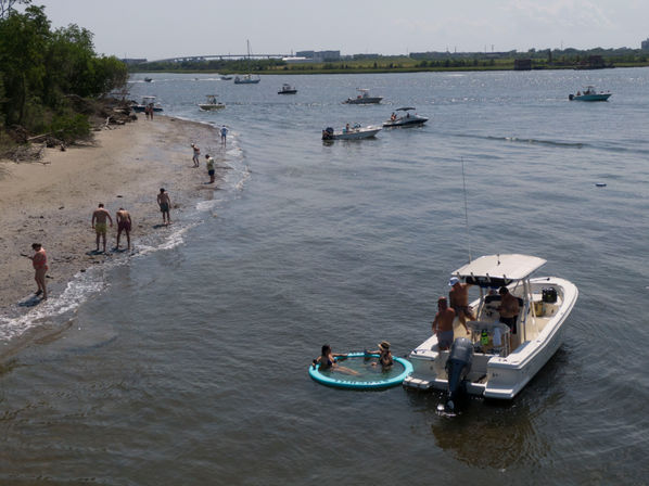 Summer bay scene with beachgoers along a sandy shoreline and multiple motorboats offshore; friends socializing on a white powerboat with a round turquoise inflatable raft beside it.