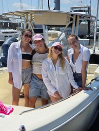 Four friends smiling and posing on a sunlit motorboat at a busy marina, wearing summer shirts, caps and sunglasses with yachts and docks in the background.