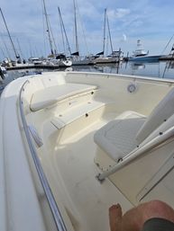 Cozy bow seating of a white motorboat docked in a calm marina, with sailboats and yachts reflected on the water under a blue, partly-cloudy sky.