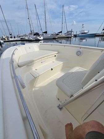 Cozy bow seating of a white motorboat docked in a calm marina, with sailboats and yachts reflected on the water under a blue, partly-cloudy sky.