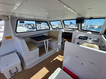 Bright boat cabin interior with cushioned dinette and table, helm station with steering wheel and electronics, cooler on deck, and wide windows looking out at sailboats and docks in a sunny marina.