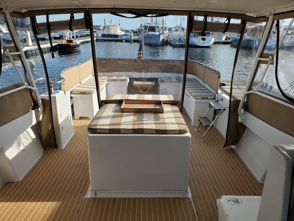 Covered boat aft deck with striped cushioned sunpad and wooden tray, faux-teak flooring and clear vinyl curtains framing a marina view of docked yachts and an American flag