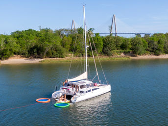 White catamaran anchored off a tree-lined shore on a sunny day with a cable-stayed bridge in the background, people relaxing on deck and colorful inflatable water rings tied alongside on calm blue water.