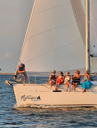 Group of people lounging on the bow of a white sailboat with a tall sail, cruising calm coastal waters in golden sunset light.