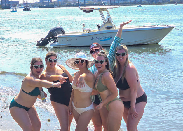 Six friends in bikinis and heart-shaped sunglasses posing cheerfully on a sunny beach shoreline with a white motorboat anchored in the coastal bay behind them