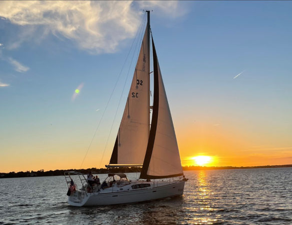 White sailboat with tall mainsail gliding across calm water at golden sunset, sun reflecting on ripples and a few people on deck enjoying the view.