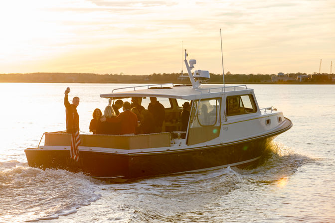 Friends on a small cabin motorboat cruising at sunset, a person waving from the stern beside an American flag, warm golden light over calm coastal waters and a distant shoreline.