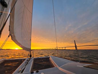 Sailboat deck and raised white sail at golden-hour sunset, looking across choppy water to a cable-stayed bridge on the horizon with warm orange and blue sky.