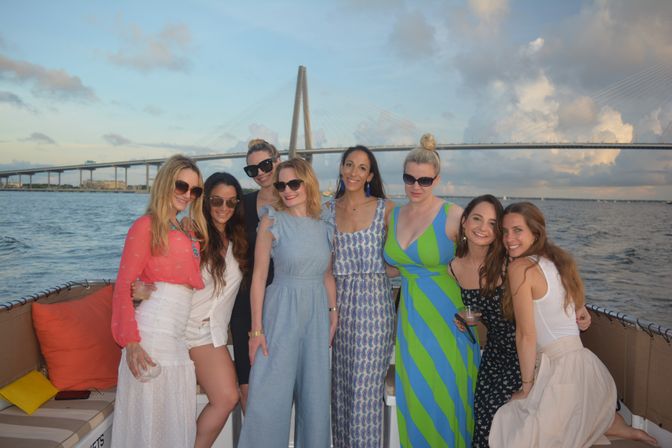 Eight women posing on a sunset boat cruise in Tampa Bay, smiling on the deck with a cable-stayed bridge and calm water in the background.