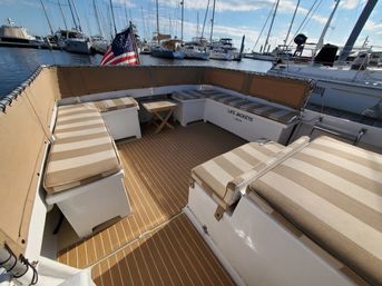 Sunlit boat cockpit with teak-style deck, striped bench cushions, life-jacket storage and an American flag, docked at a marina with sailboats in the background.