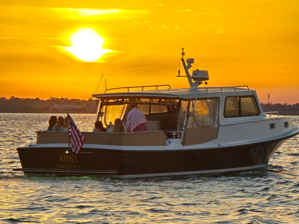 Motorboat with passengers cruising on calm coastal waters at a golden sunset, American flag fluttering and shoreline silhouette in the background.