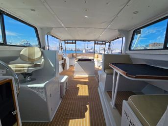 Bright yacht cabin interior with teak-style flooring, helm chair and dinette table, panoramic windows framing a sunny marina with moored boats