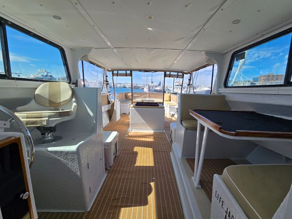 Bright yacht cabin interior with teak-style flooring, helm chair and dinette table, panoramic windows framing a sunny marina with moored boats
