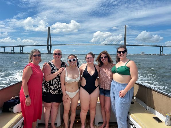 Six women enjoying a sunny boat outing in swimsuits with a cable-stayed bay bridge and blue sky backdrop