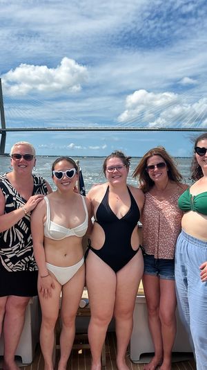 Six women enjoying a sunny boat outing in swimsuits with a cable-stayed bay bridge and blue sky backdrop