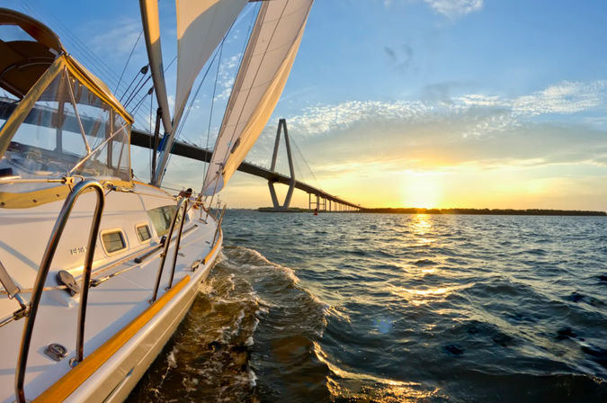 Sailboat with full sails gliding under a cable-stayed bridge at golden sunset, sun reflecting on choppy harbor waters