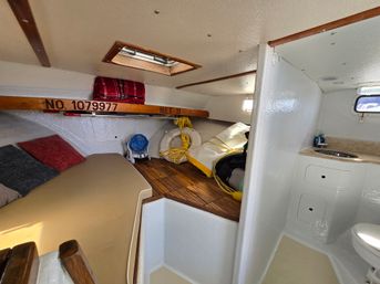 Cozy sailboat cabin interior with v-berth cushions, teak plank floor, overhead hatch, lifesaver ring and coiled ropes, small sink and marine head, and compact storage with wood trim.